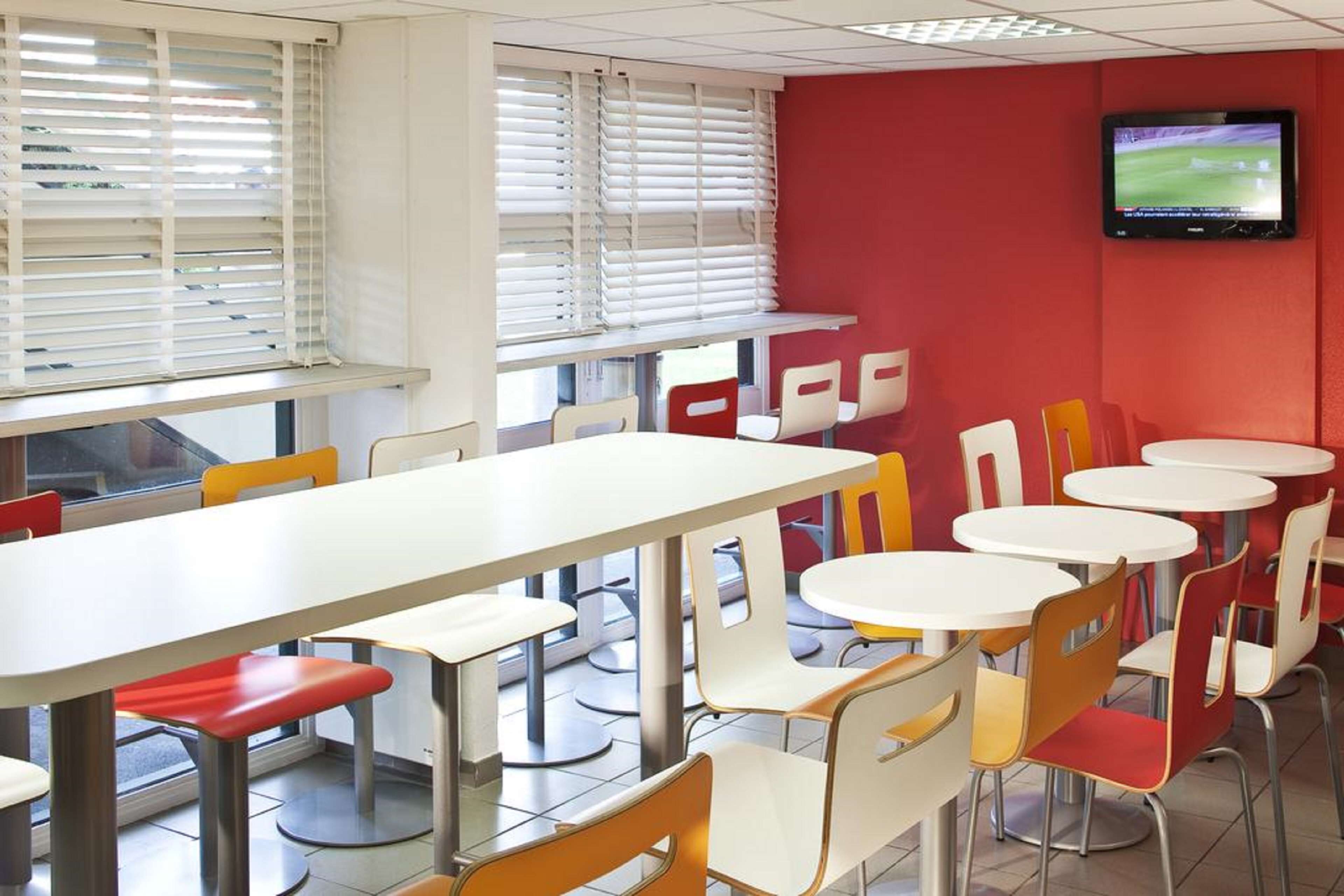 A dining area with tables and chairs, a television mounted on the wall, and windows with blinds.