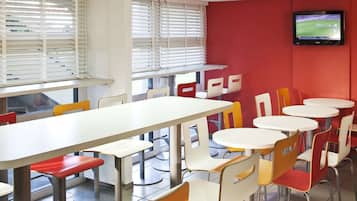 Un espace repas avec des tables et des chaises, un téléviseur fixé au mur et des fenêtres équipées de stores.