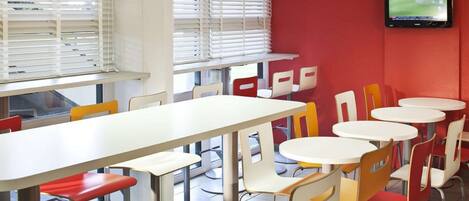 A dining area with tables and chairs, a television mounted on the wall, and windows with blinds.