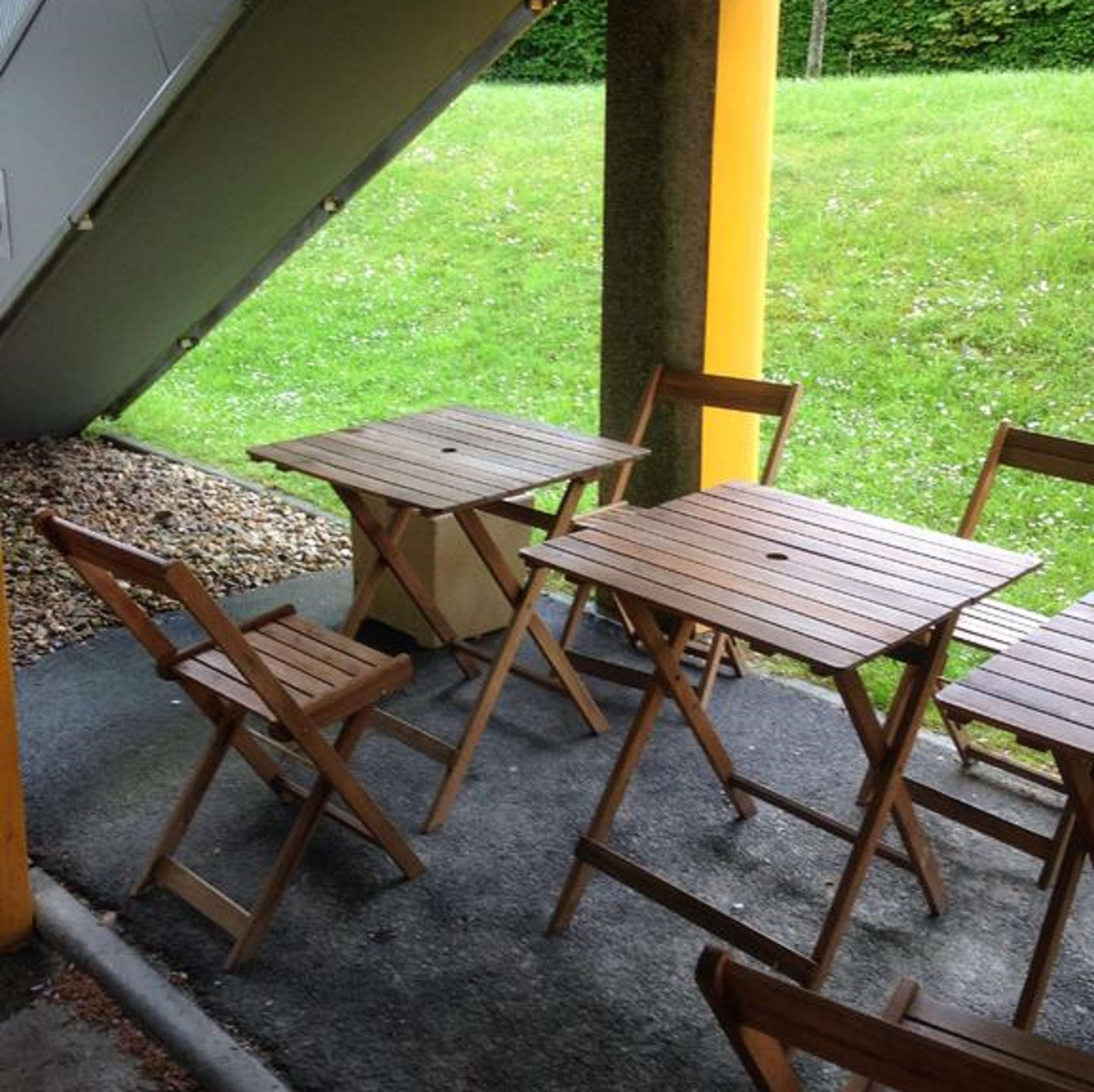 Un patio con mesas y sillas de madera, suelo de concreto y un césped al fondo.