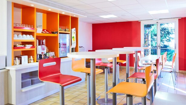 A brightly lit cafeteria with orange and white furniture, a counter with various items, and a view of greenery outside. - Premiere Classe Charleville Mezieres (Charleville-Mezieres)