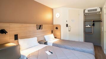 Ein Hotelzimmer mit zwei Betten, einem Holzkopfteil, Nachttischlampen, einer Wanduhr, einer Tür mit Handtuchhalter und einer Klimaanlage.