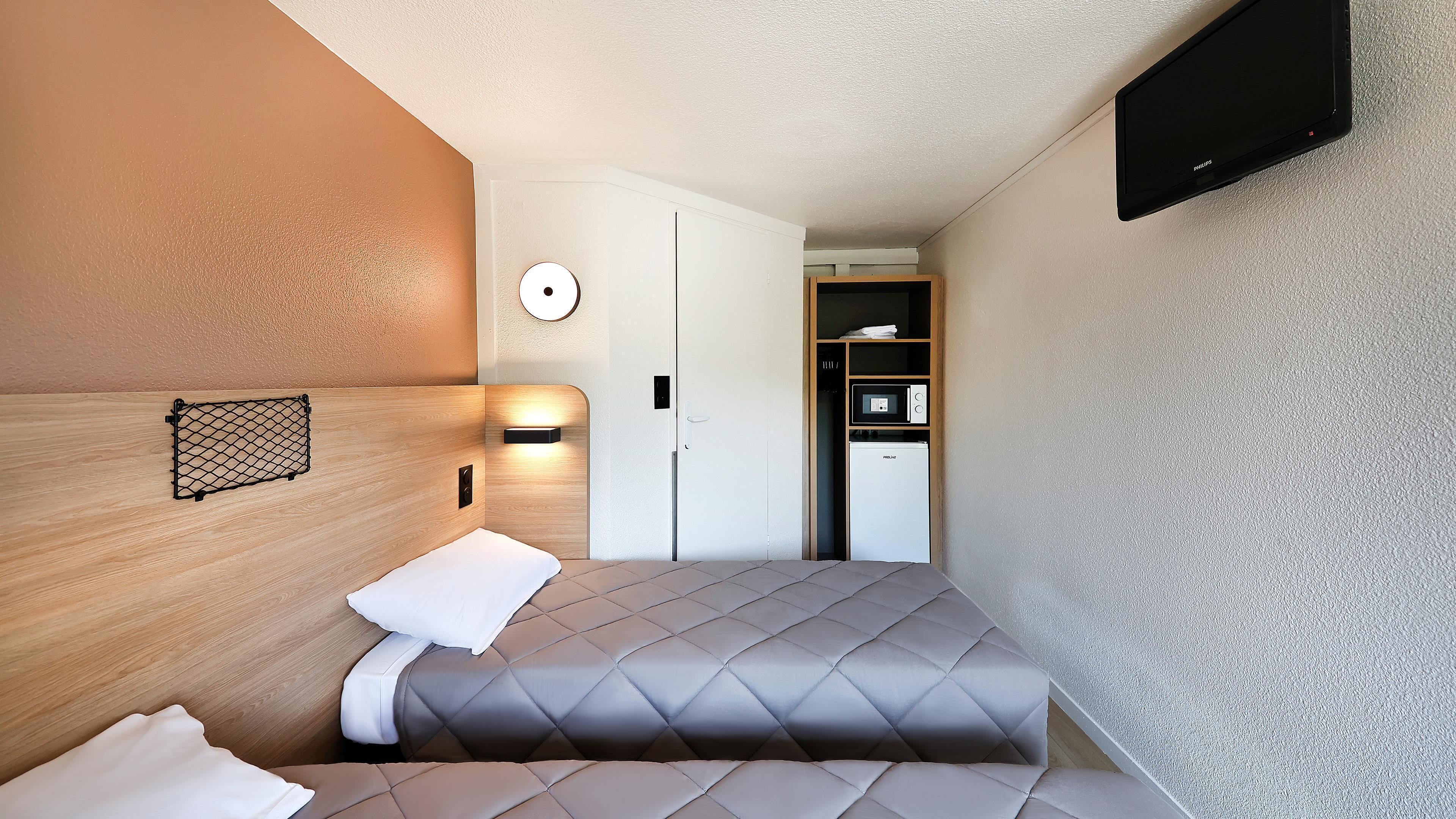 A compact hotel room with a bed, a bedside table, a wall-mounted TV, a microwave, and a small kitchenette.