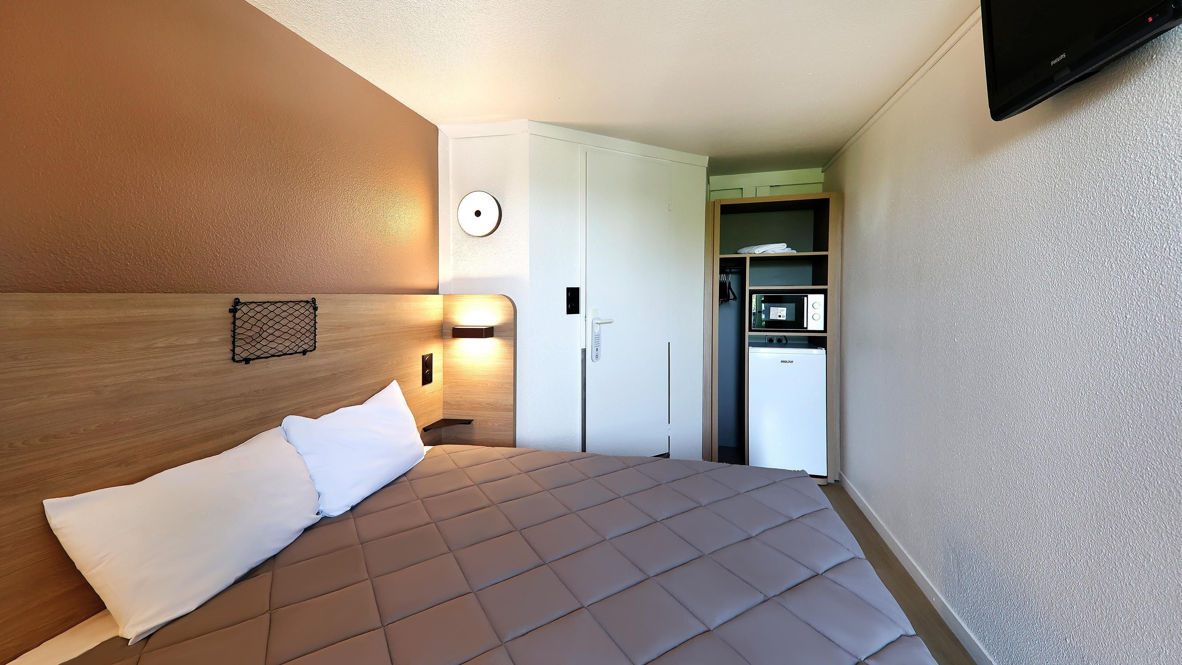 Una habitación de hotel con una cama, un televisor montado en la pared, una puerta con manija y un refrigerador pequeño.