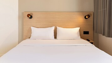 Una cama bien tendida con dos almohadas, un cabecero de madera con dos luces fijadas a la pared, y una mesita de noche con un control remoto.