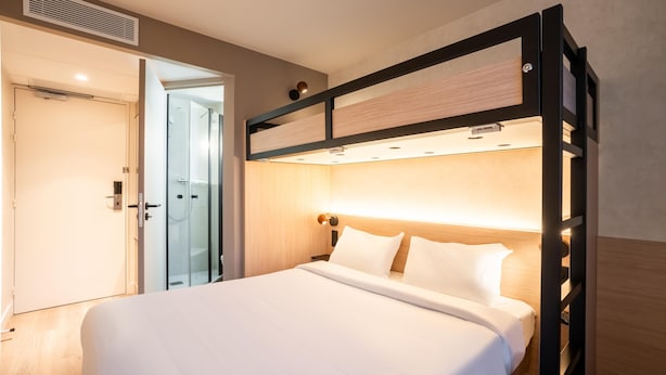 Quarto de hotel moderno com cama, mesa de cabeceira, luminária fixada na parede e porta de acesso a um banheiro.