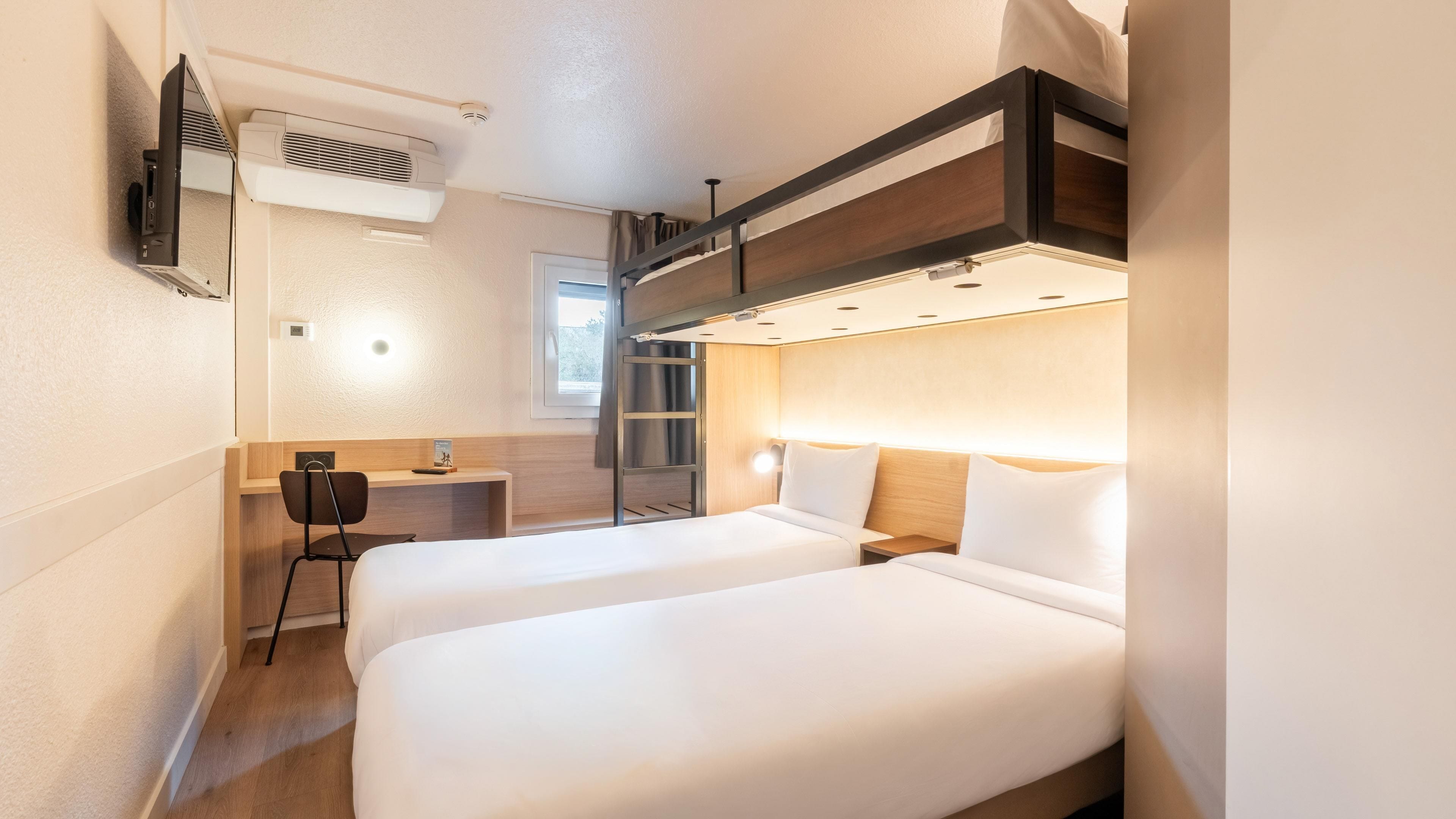 A hotel room with two beds, a desk, a chair, a TV, and a window.
