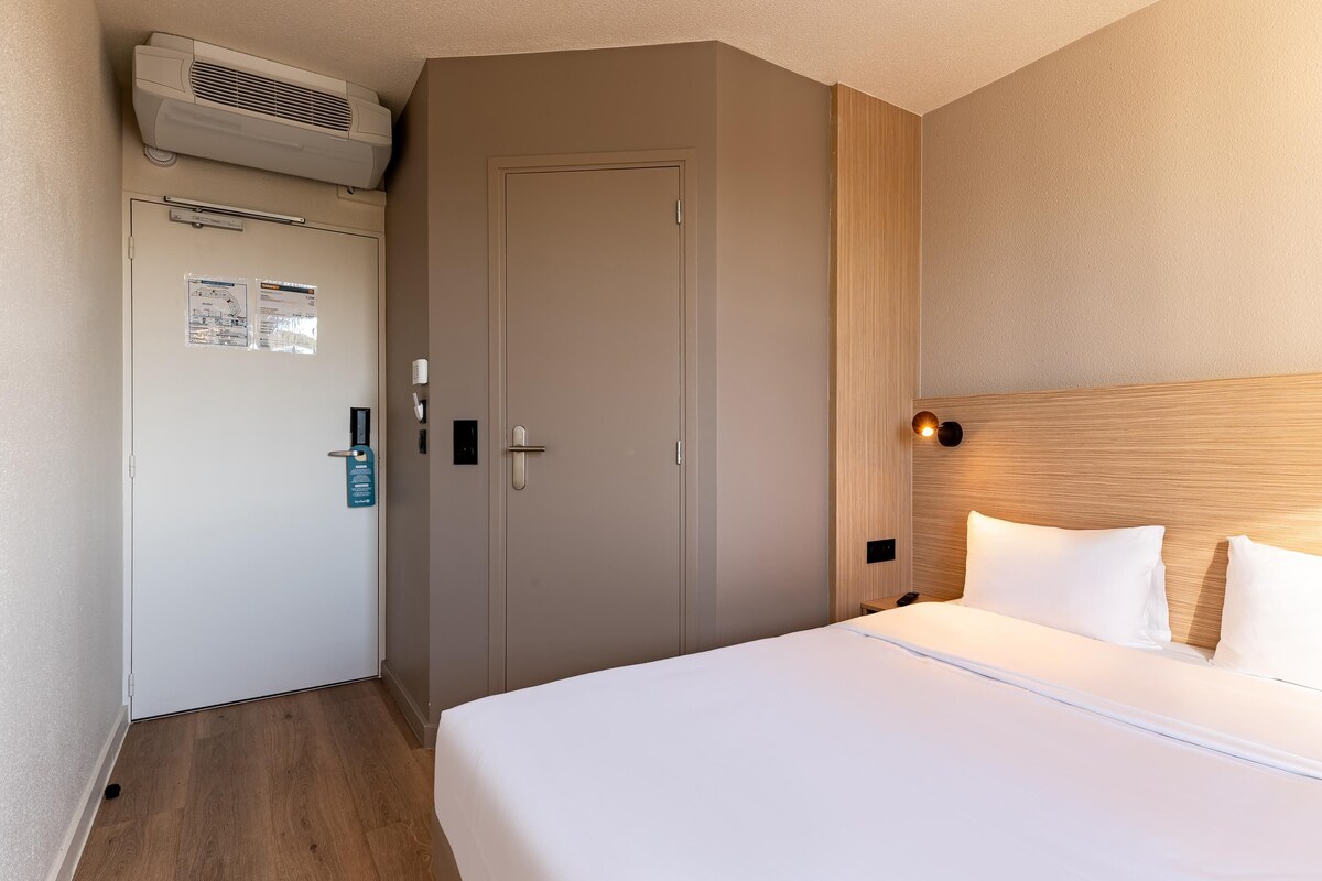 Hotellihuone, jossa on sänky, ovi, ilmastointilaite ja seinään kiinnitetty valaisin.