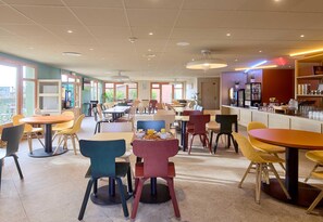 A spacious dining area with multiple tables and chairs, a bar counter, and large windows allowing natural light. - B&B HOTEL Saint-Witz Roissy (Saint-Witz)