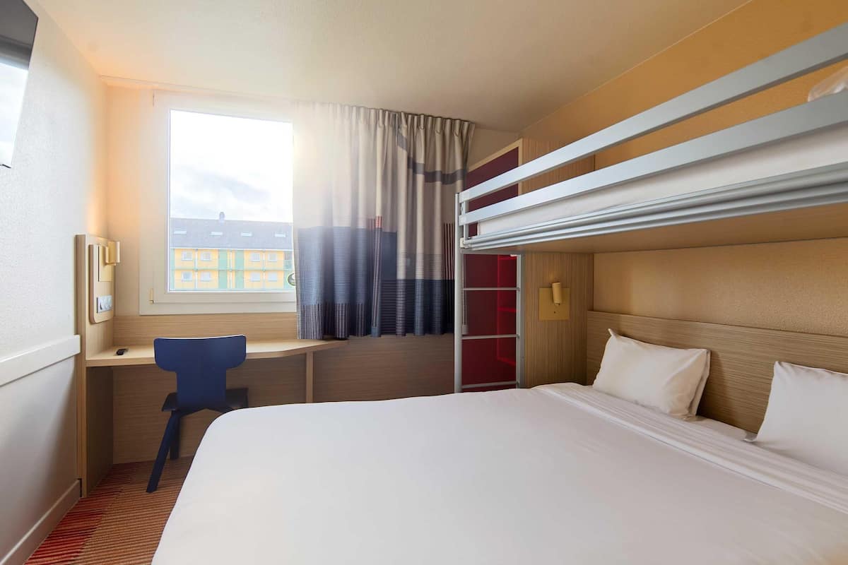 A hotel room with a bunk bed, a desk with a chair, a window with curtains, and a view of buildings outside.