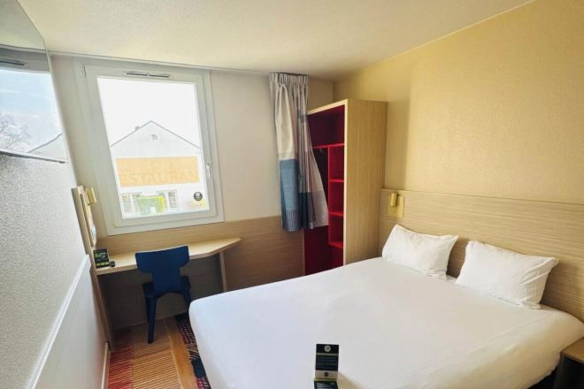 A hotel room with a bed, a desk, and a chair. There is a window with a view of buildings outside.