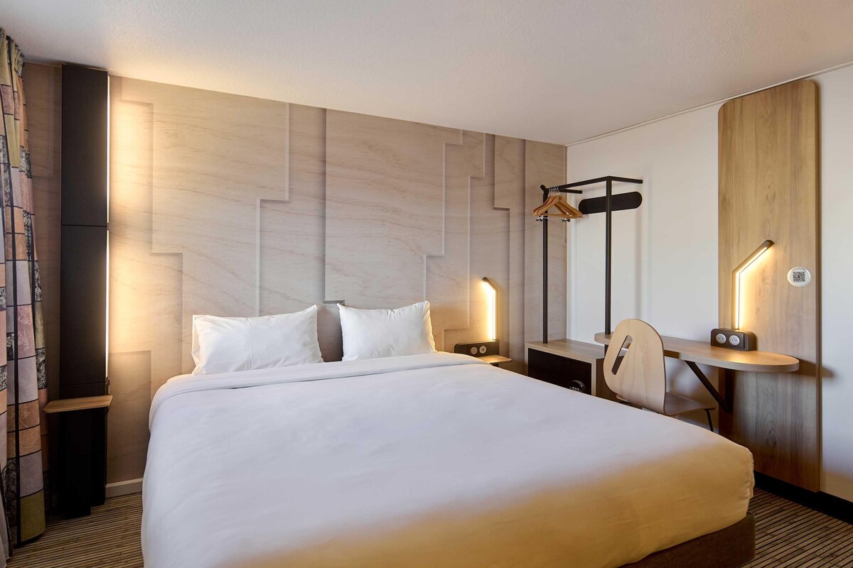 Ein Hotelzimmer mit einem großen Bett, Nachttischen, einem Schreibtisch mit Stuhl, einem Spiegel und einem Kleiderhaken.