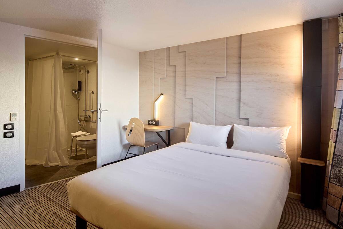 A hotel room with a large bed, a desk with a chair, a small table with a lamp, a shower area, and a wooden headboard.