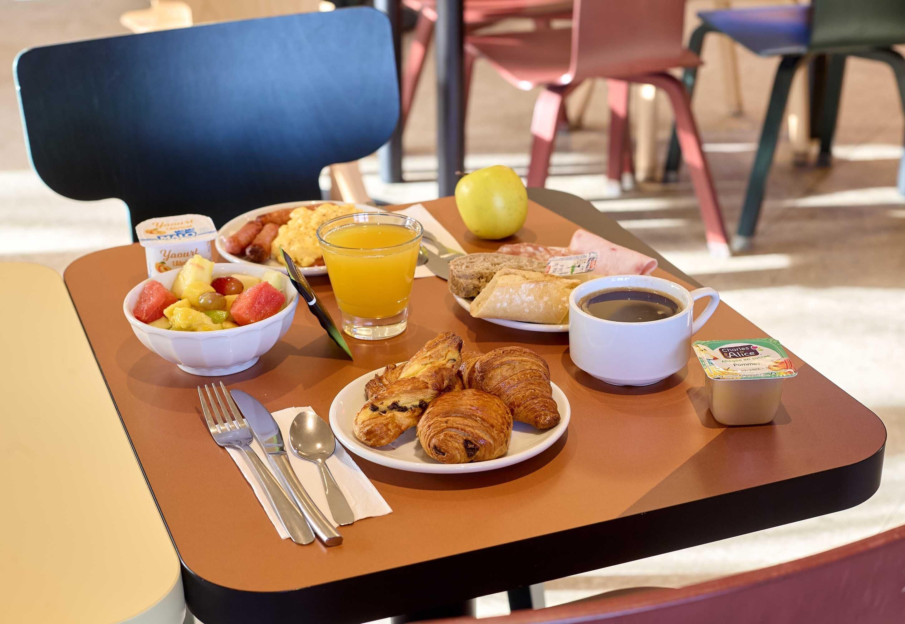 Een ontbijttafel met croissants, fruit, sap, koffie en een yoghurtbeker.