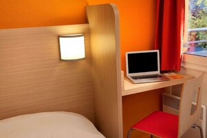 Une chambre d’hôtel de taille réduite, comprenant un lit, un bureau avec un ordinateur portable, une chaise rouge et une lampe fixée au mur.