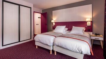 Quarto de hotel com cama de casal, duas mesas de cabeceira, um criado-mudo, uma escrivaninha, uma cadeira, um guarda-roupa e uma porta.