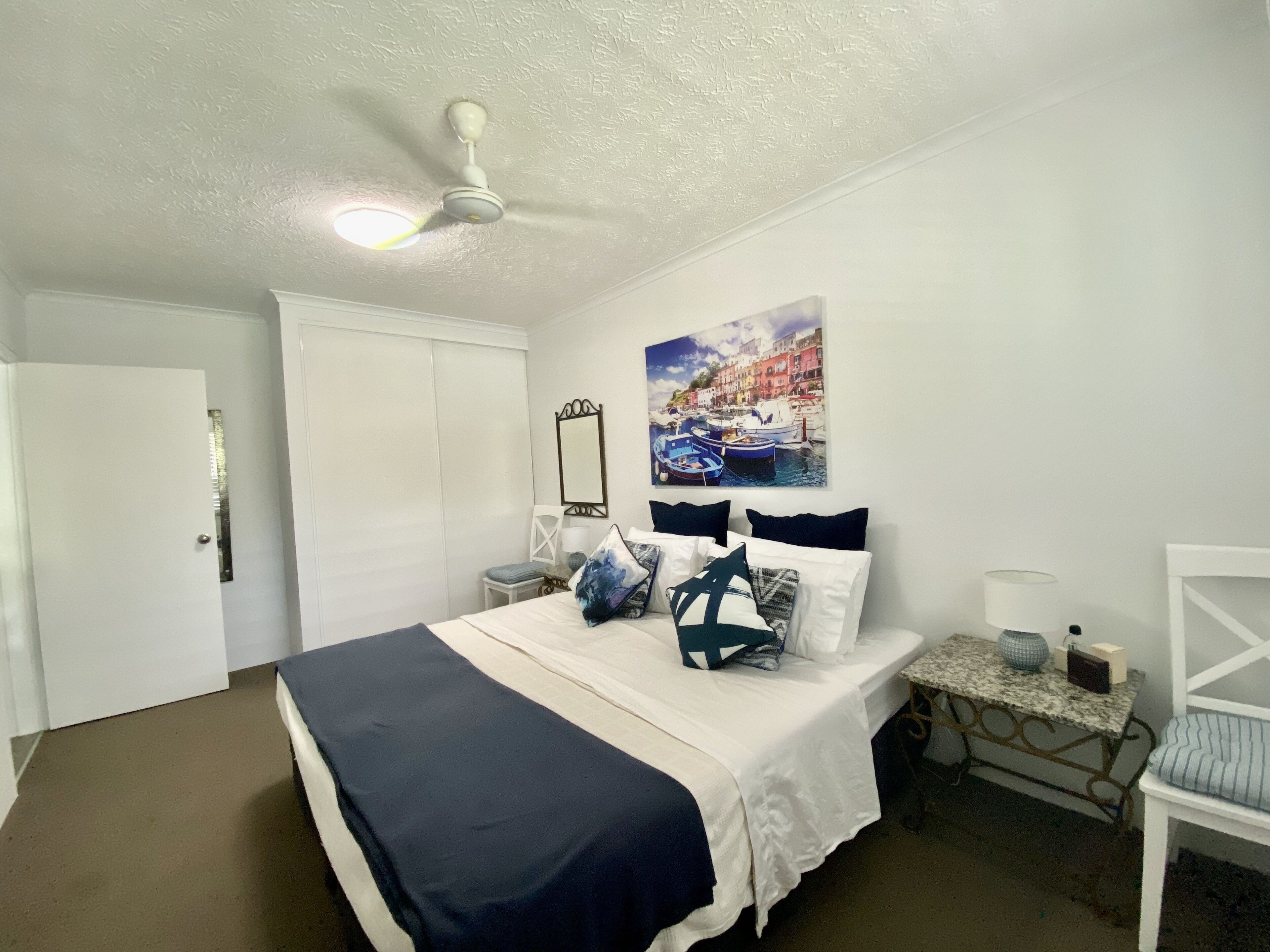 Un dormitorio con una cama, una mesita de noche, una silla, un ventilador de techo y un espejo instalado en la pared.