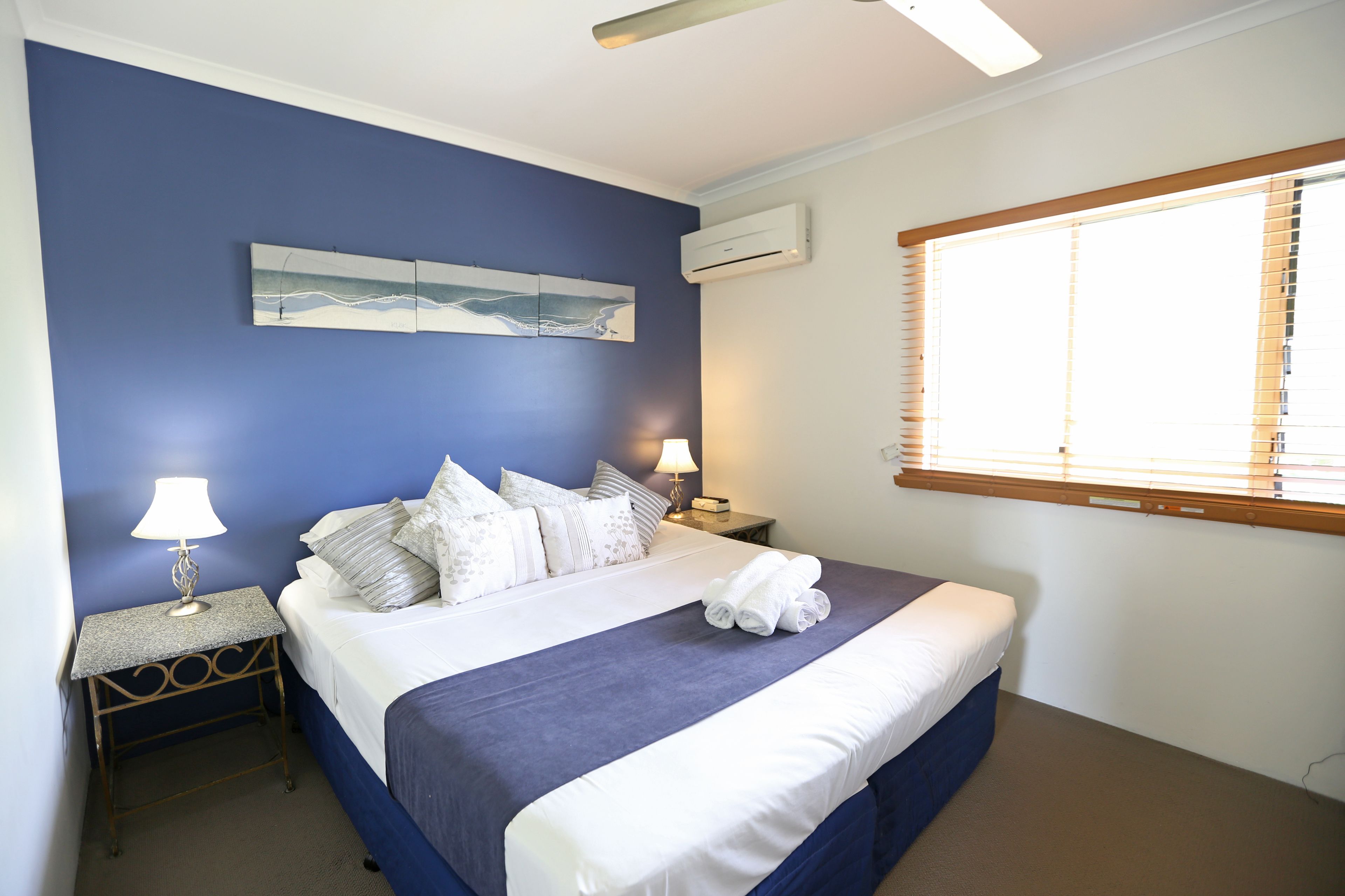 Un dormitorio con cama, dos mesitas de noche, aire acondicionado instalado en la pared y ventana con persianas.