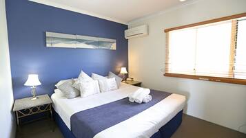 Un dormitorio con cama, dos mesitas de noche, aire acondicionado instalado en la pared y ventana con persianas.