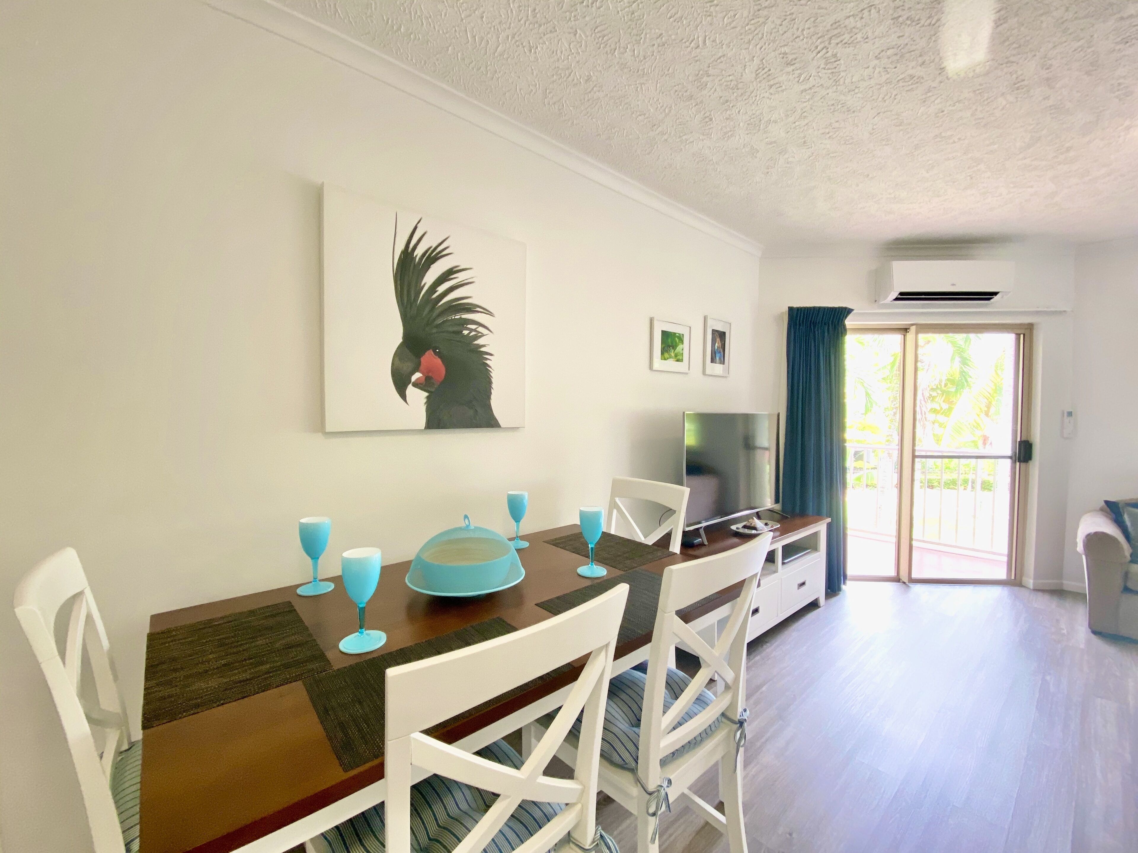 Un comedor con una mesa de madera, sillas blancas y una camino de mesa azul. Hay un cuadro de un pájaro en la pared y un televisor sobre un mueble.