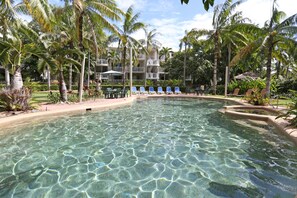 A clear pool surrounded by palm trees and lounge chairs, with a building in the background.