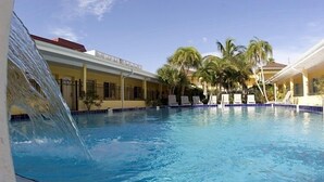 A swimming pool with a waterfall feature, surrounded by buildings with balconies and palm trees.
