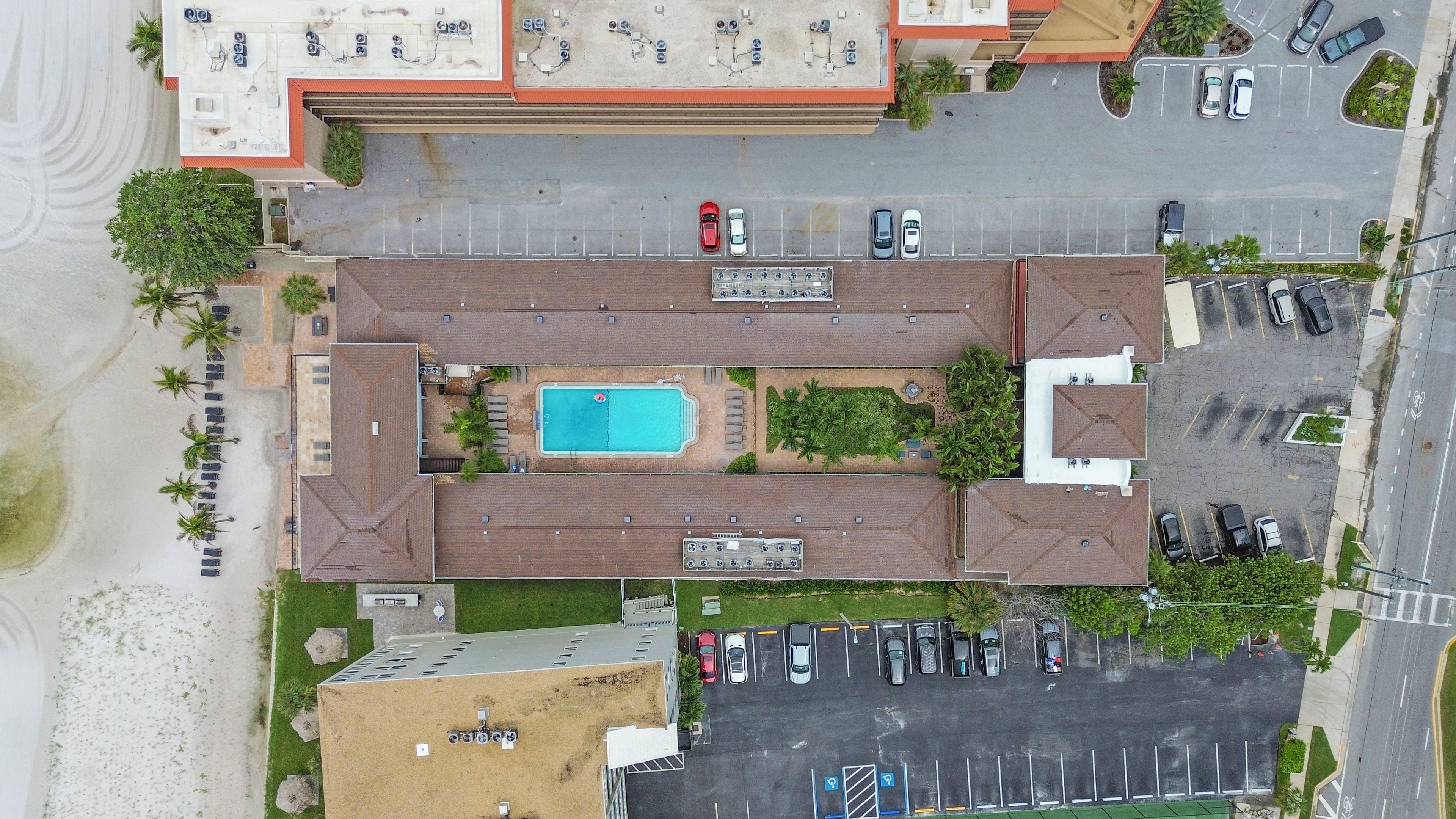 An aerial view of a building with a swimming pool, parking lot, and adjacent beach.