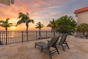 A balcony with a view of the sunset over the ocean, palm trees, and a building with a red roof. - Hotel Sol (Redington Shores)