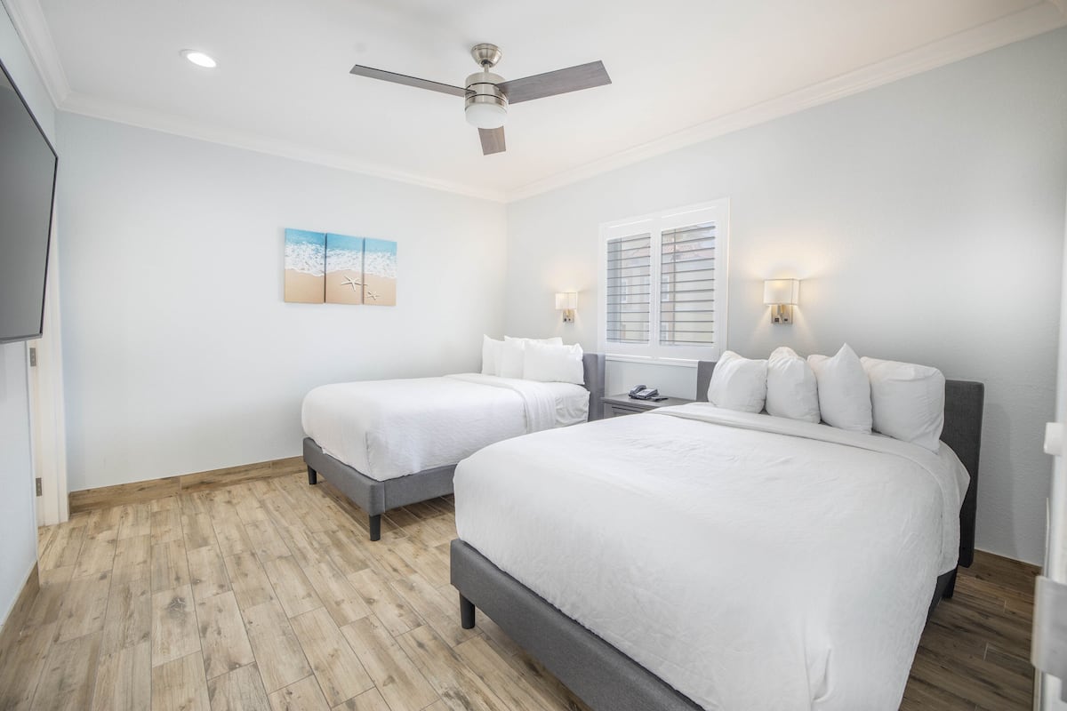 A bedroom with two beds, a ceiling fan, a window with blinds, and a painting on the wall.