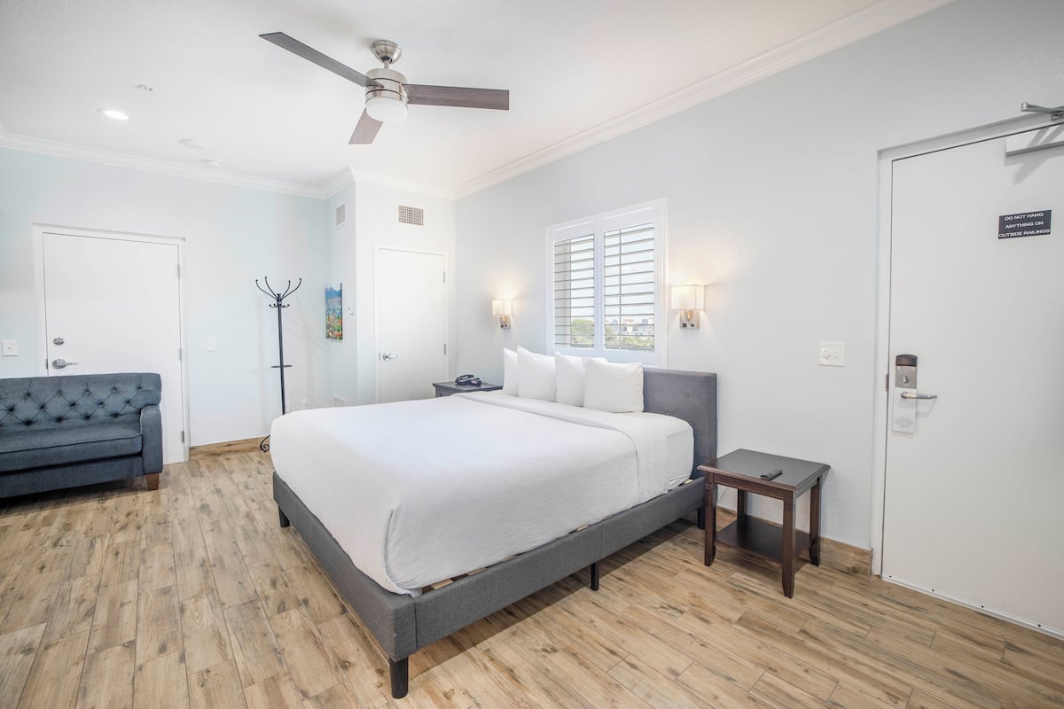A bedroom with a large bed, a ceiling fan, a nightstand, a small table, a sofa, and a door with a sign.