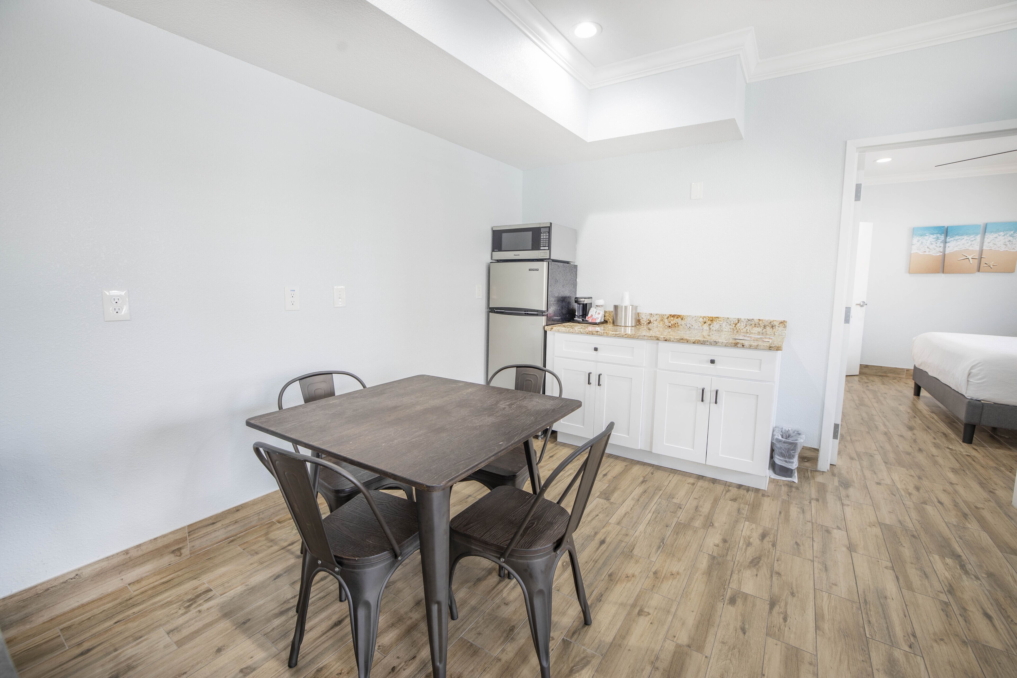 A modern kitchen with white cabinets, a built-in microwave, and a coffee machine. There is a dining area with a wooden table and metal chairs.