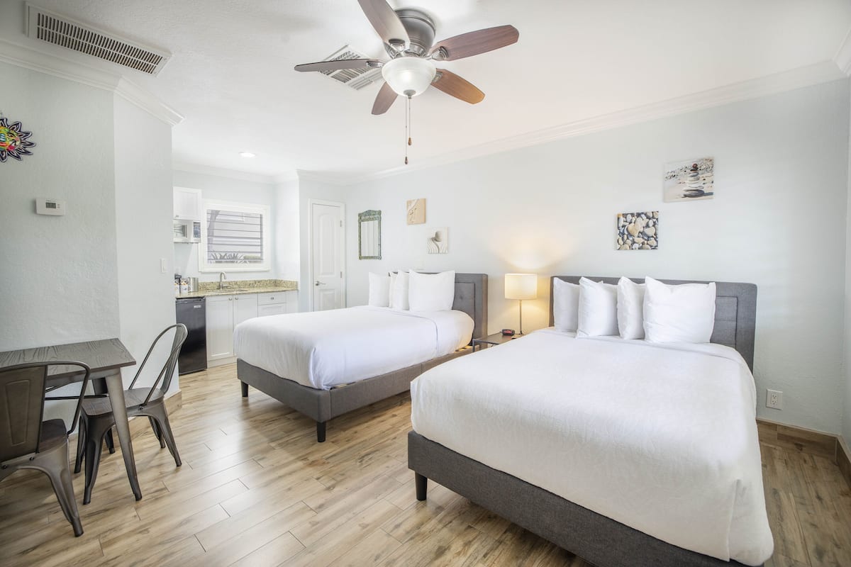A hotel room with two beds, a desk, chairs, a ceiling fan, and artwork on the walls.