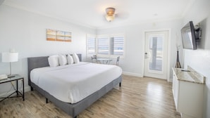 A bedroom with a large bed, a bedside table, a desk, and a chair. There is a window with blinds, a door, and a ceiling light fixture.