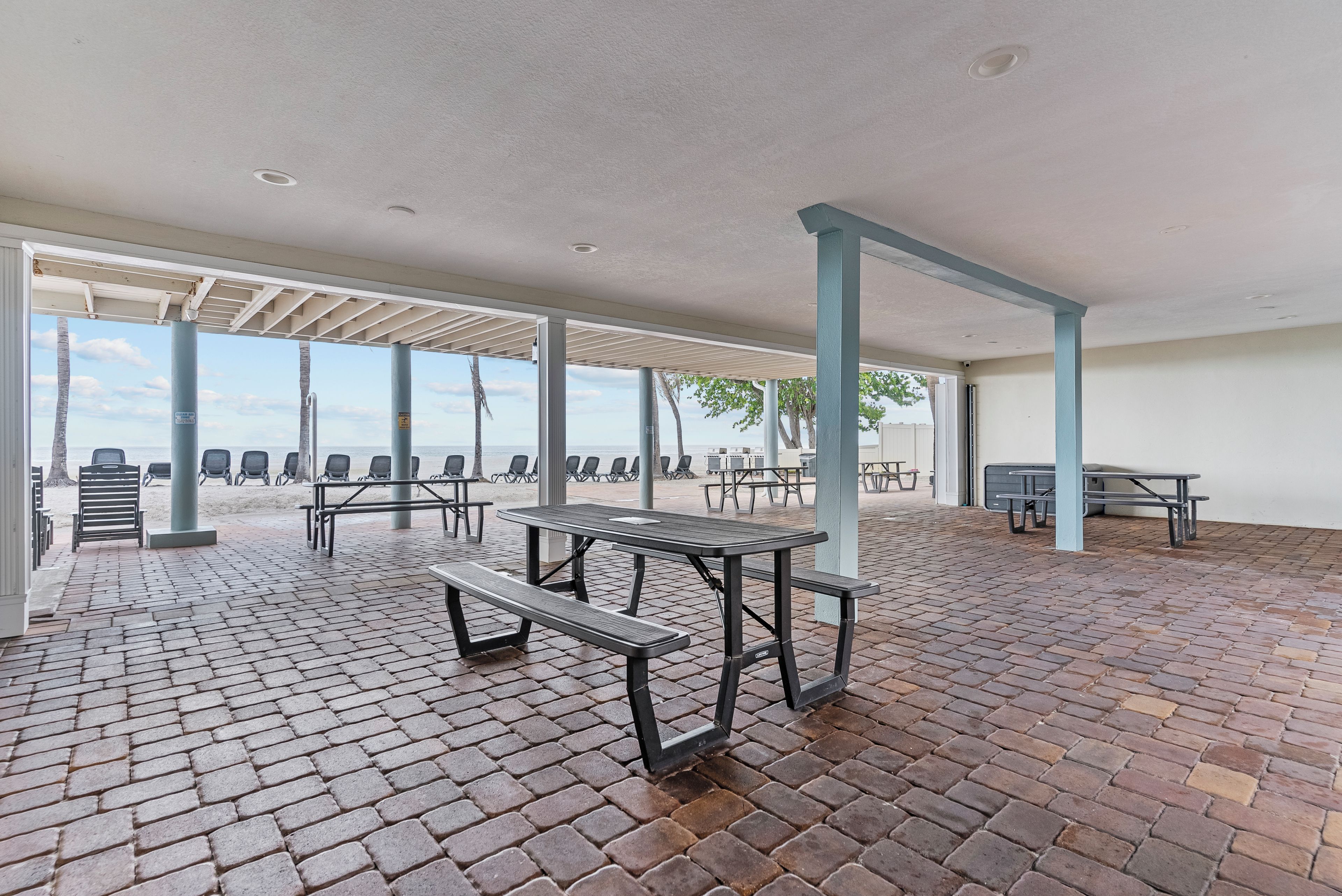 An outdoor patio area with picnic tables and chairs, a brick floor, and a view of the beach.