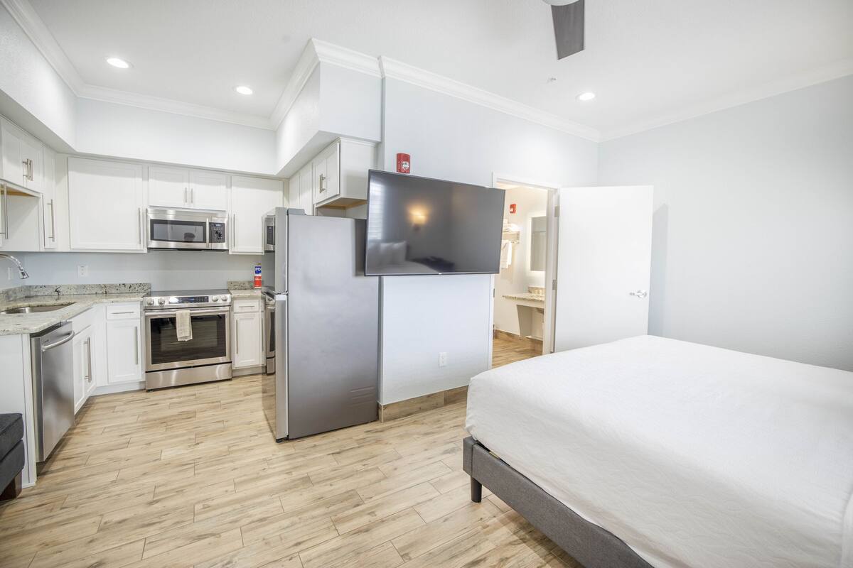 A modern hotel room with a large bed, a kitchenette with a microwave, oven, and refrigerator, and a flat-screen TV mounted on the wall.