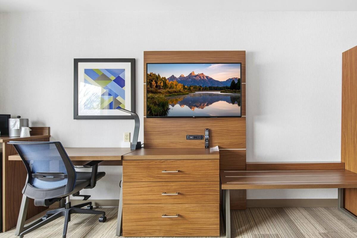 Hotelkamer met een houten bureau, een stoel, een flatscreen televisie aan de muur en een lijst met een berglandschap.