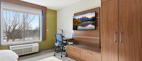 A room with a large window, a desk with a chair, a TV mounted on the wall, and wooden cabinets.