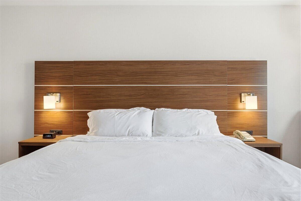 Une chambre d’hôtel avec une tête de lit en bois, deux lampes de chevet, un lit avec du linge de lit blanc et un téléphone sur la table de chevet.