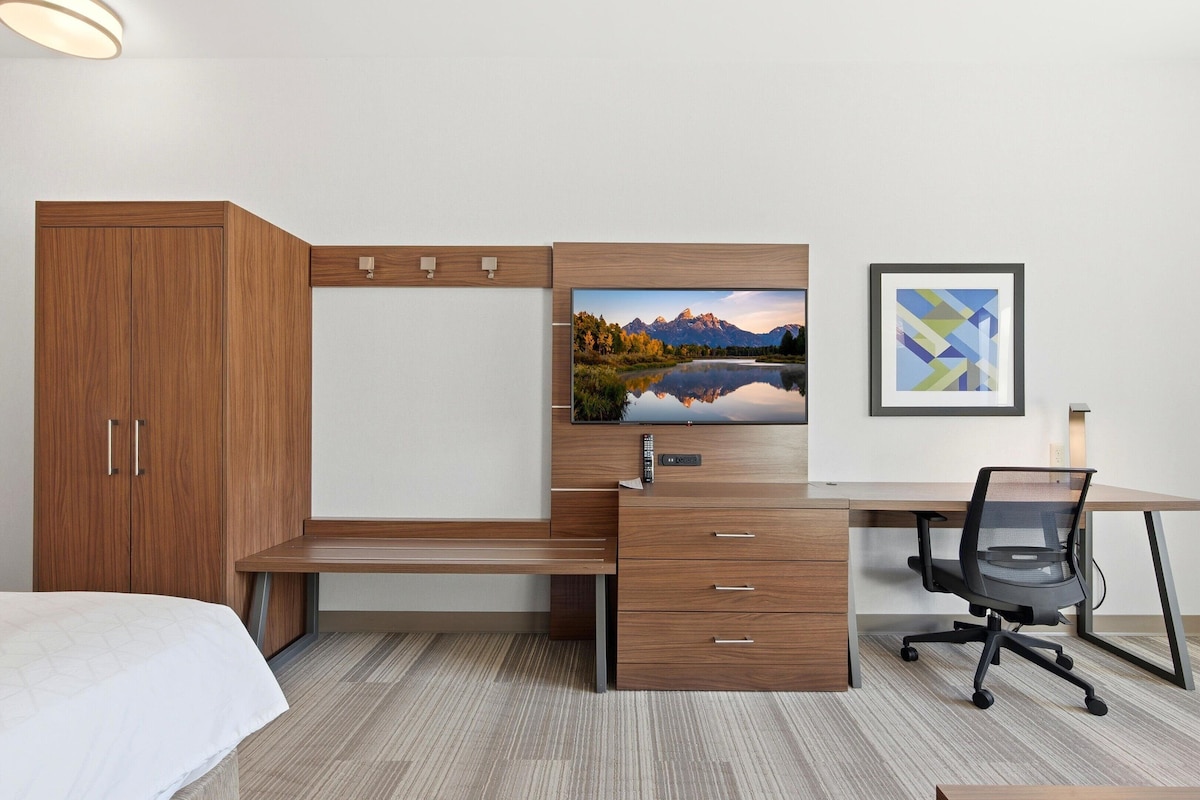 Un dormitorio con cama, armario de madera, escritorio con silla, televisor de pantalla plana y un cuadro en la pared.