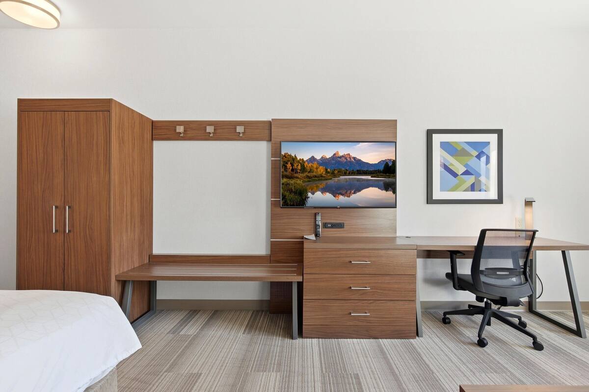 Een slaapkamer met een bed, een houten kledingkast, een bureau met stoel, een flatscreen televisie en een lijst met een afbeelding aan de muur.