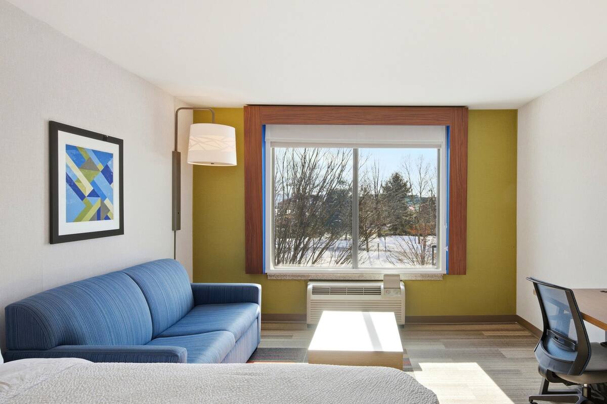 Een moderne hotelkamer met een blauwe bank, een bureau met stoel, een groot raam met uitzicht op bomen, en een lijst met abstracte kunst aan de muur.