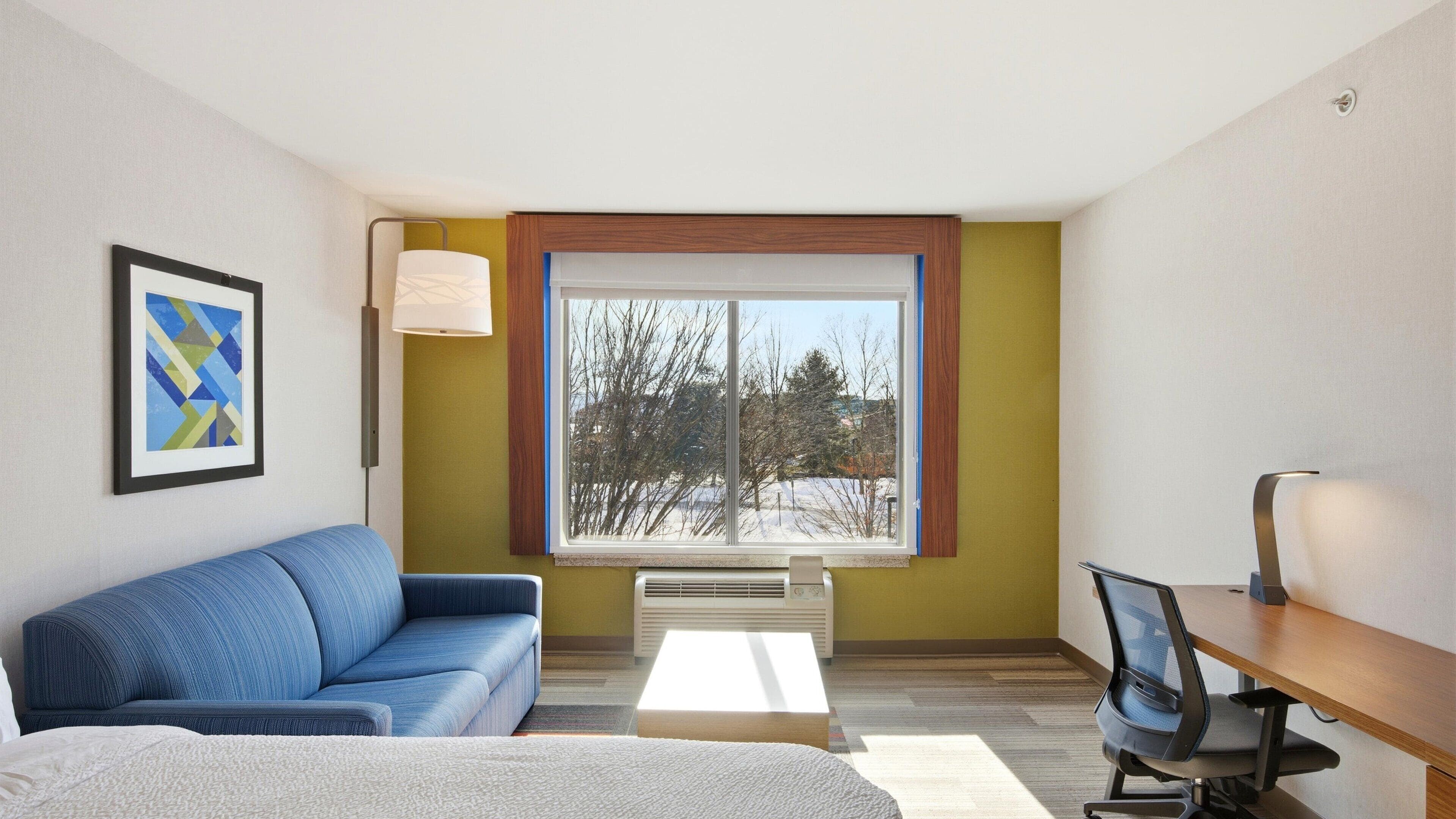 Una camera d'albergo moderna con un divano blu, una scrivania con una sedia, un'ampia finestra che si affaccia su alberi e un quadro astratto incorniciato appeso al muro.
