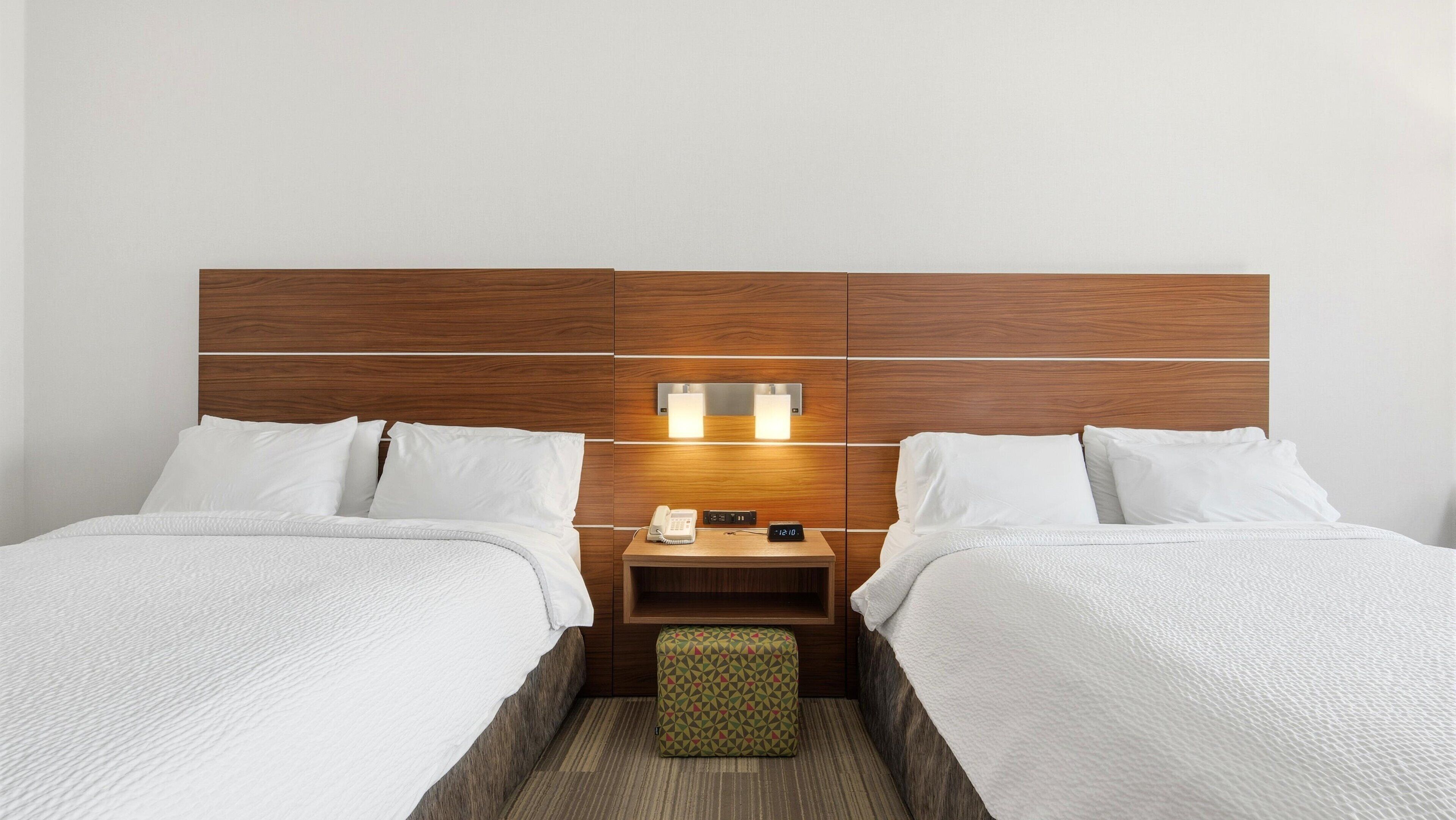 Una habitación de hotel con dos camas, un cabecero de madera, una mesita de noche con un teléfono y un reloj, y un pequeño otoman.