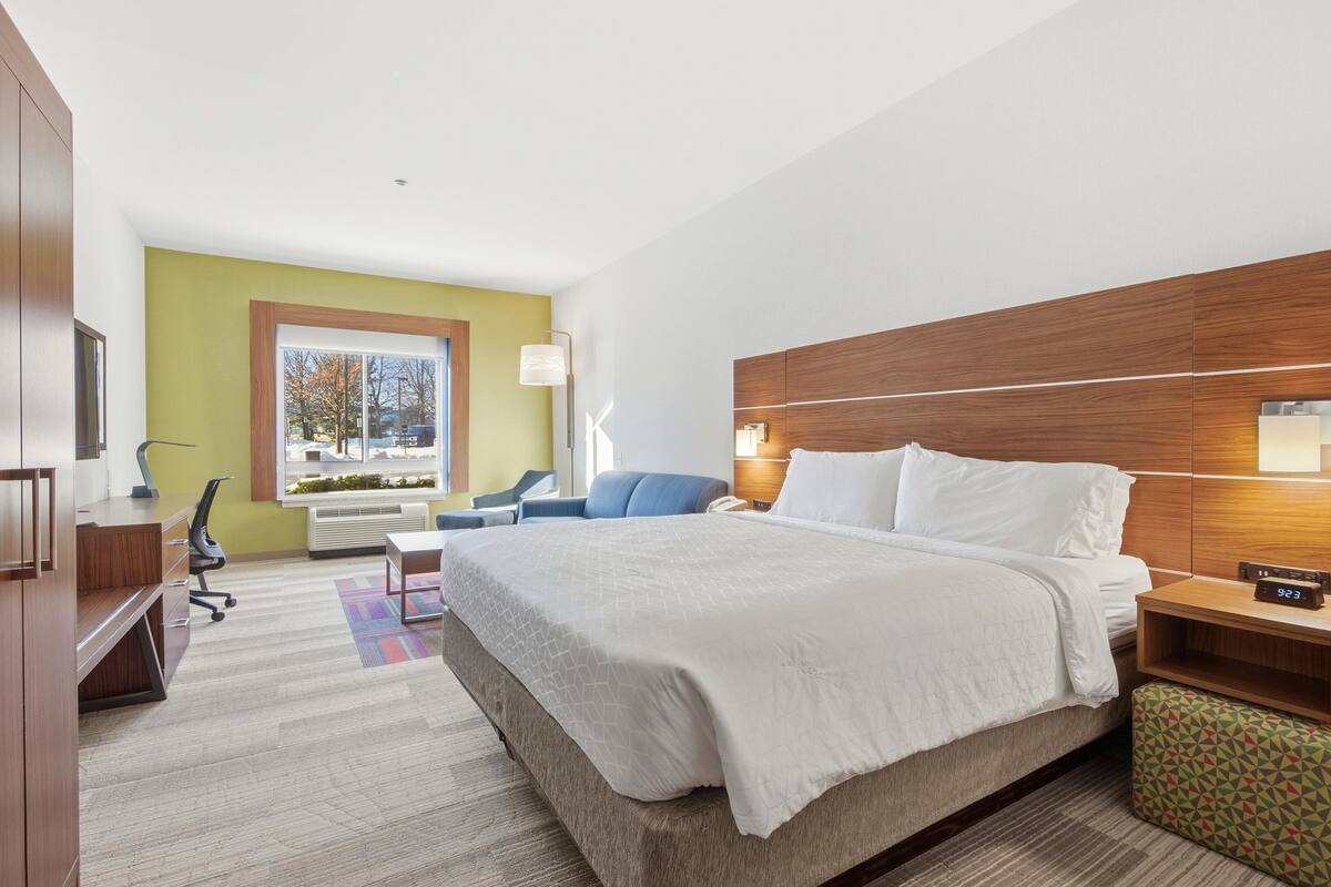 Een hotelkamer met een groot bed, een bureau, een stoel, een bank, een raam en een nachtkastje met een klok.