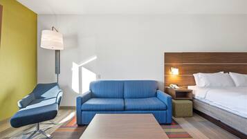 A hotel room with a blue sofa, a wooden nightstand with a lamp, a bed with white linens, and a colorful striped rug.