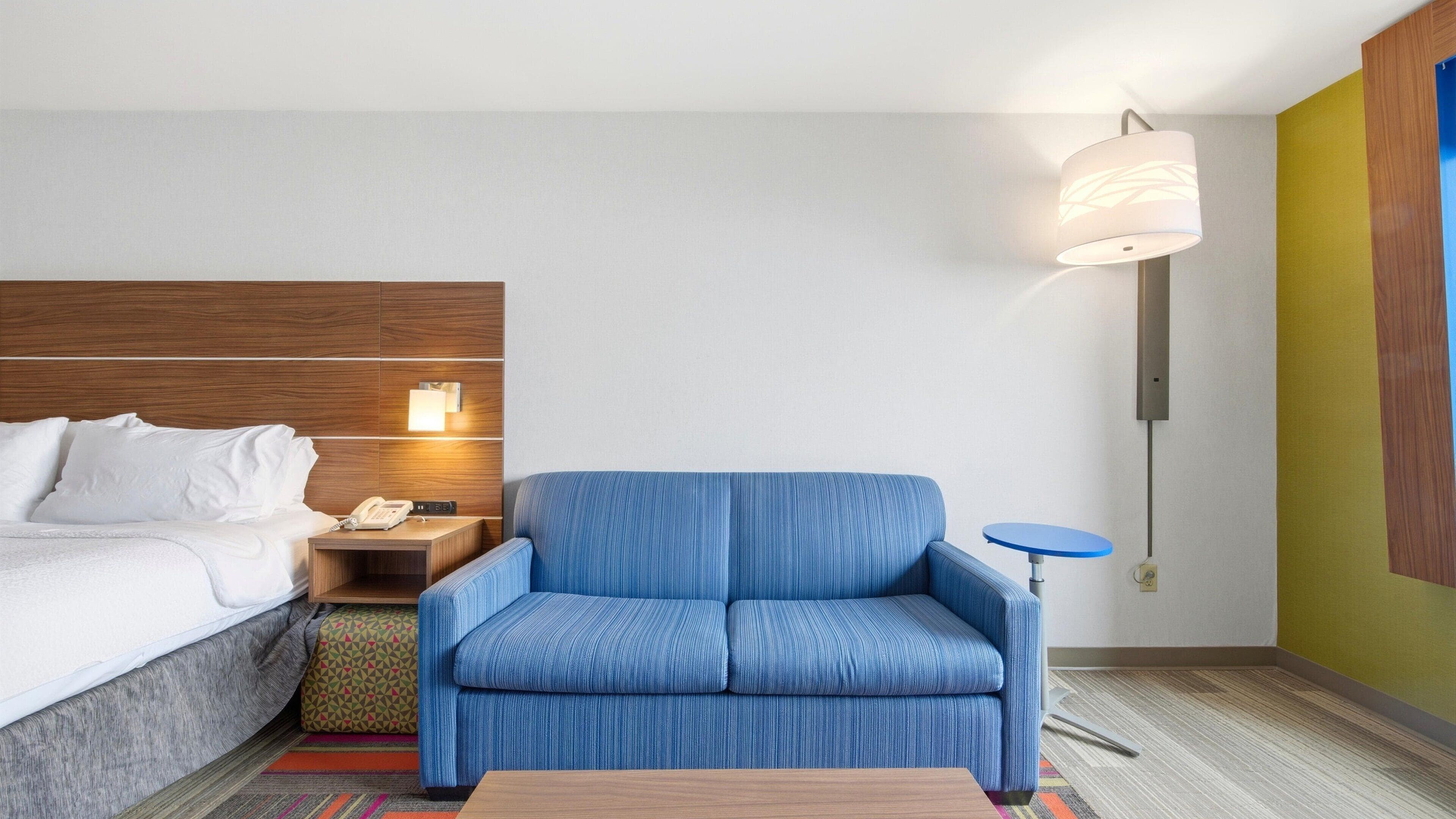 Habitación de hotel con una cama, un sofá azul, una mesita, una mesita de noche con un teléfono y una lámpara de pared.