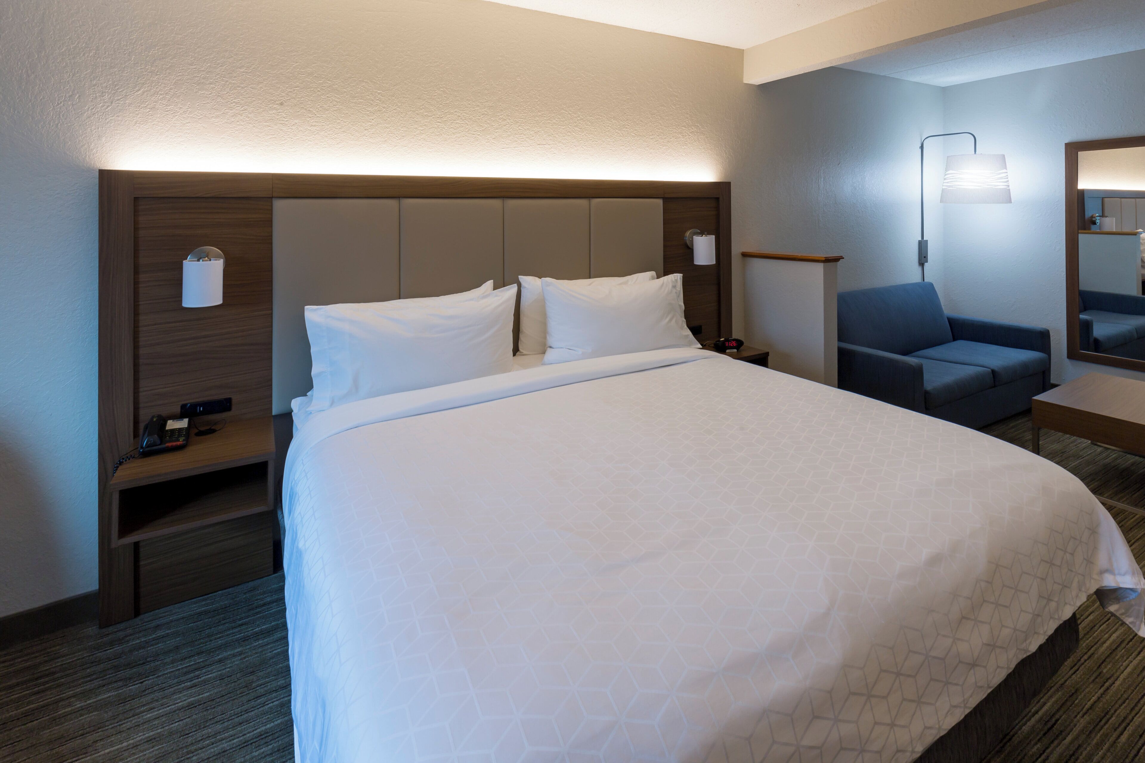 A hotel room with a large bed, a bedside table with a lamp, a chair, and a mirror.