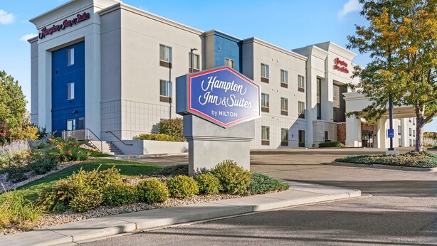 A Hampton Inn & Suites by Hilton building with a blue entrance door, a signboard, and landscaped gardens.