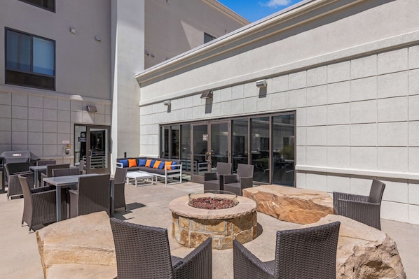 An outdoor patio area with wicker furniture, a stone fire pit, and a view of a building with large windows.