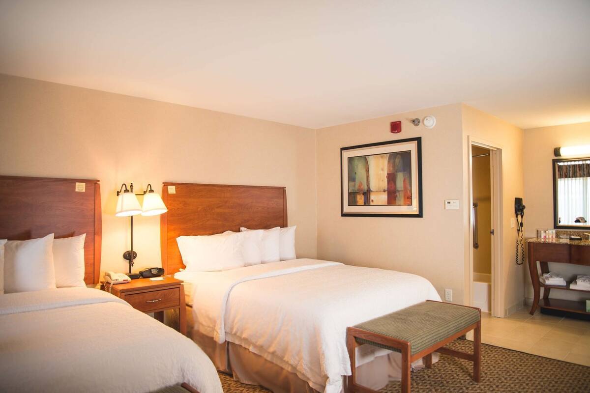 A hotel room with two beds, a bedside table with lamps, a painting on the wall, a bathroom with a mirror, and a small bench.
